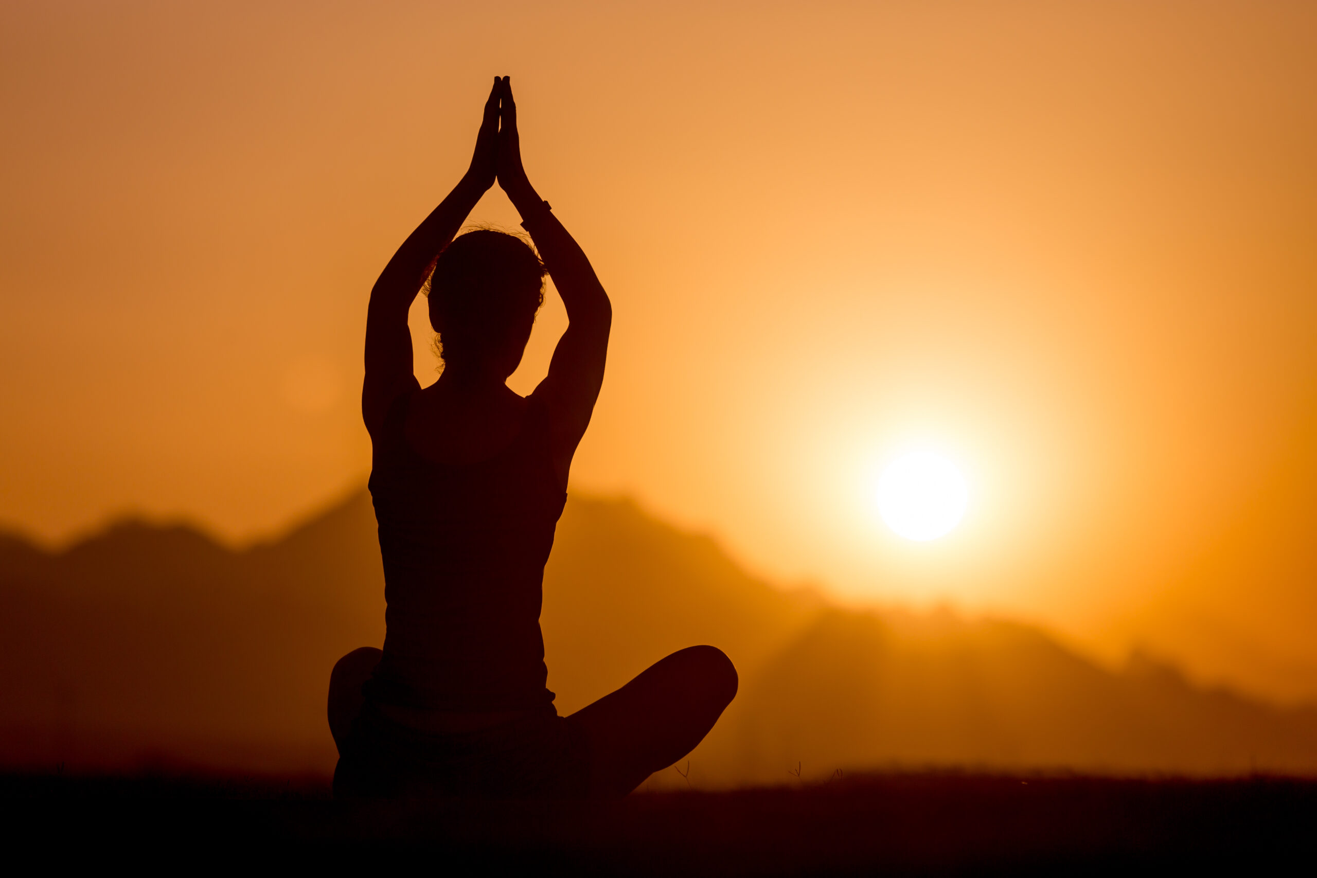 Silhouette of young woman sitting in yogic sukhasana easy posture in picturesque mountainous landscape, watching sunset or sunrise, meditating